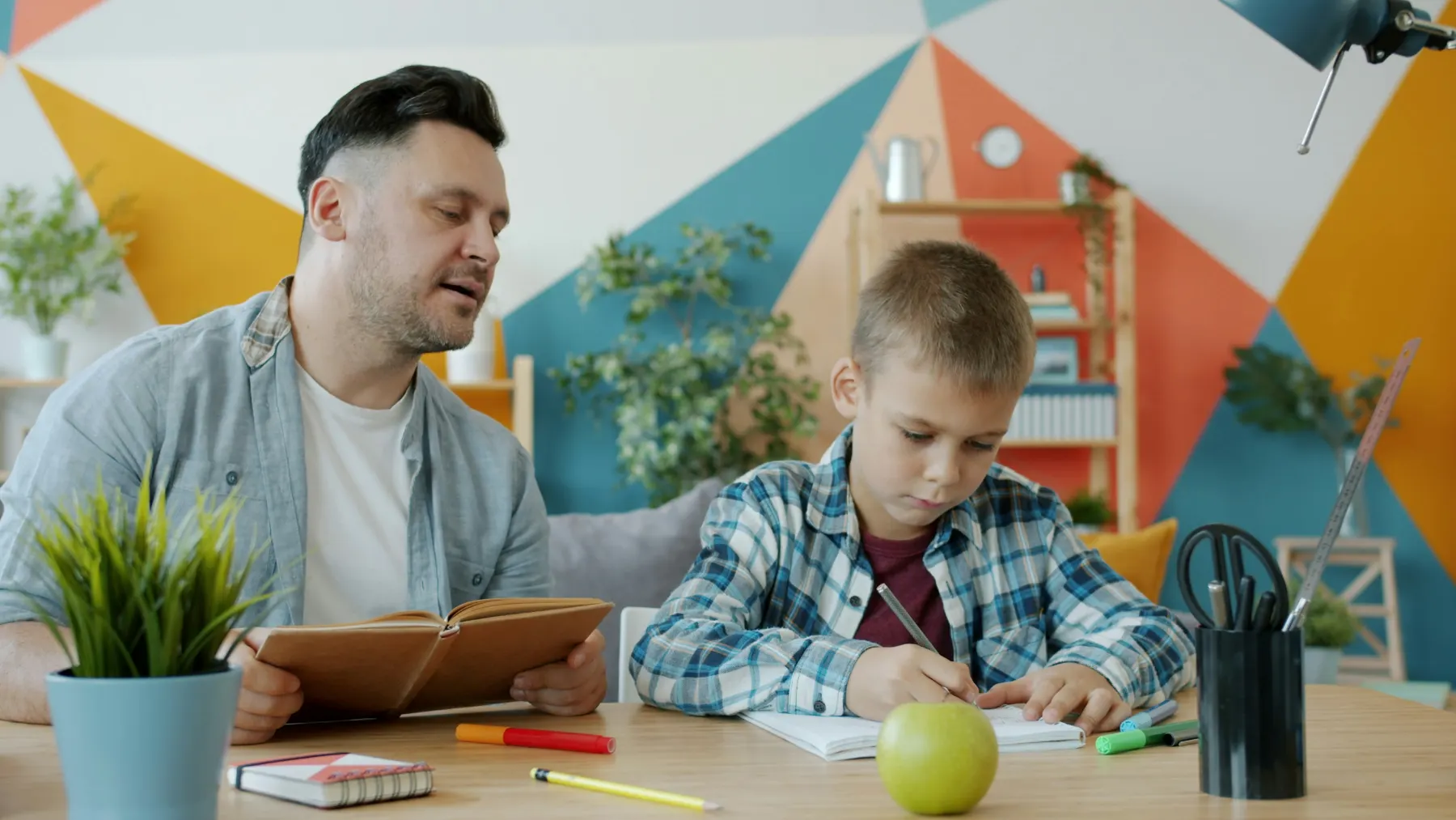 A mentor helping a child write at a desk with study supplies and a laptop nearby.